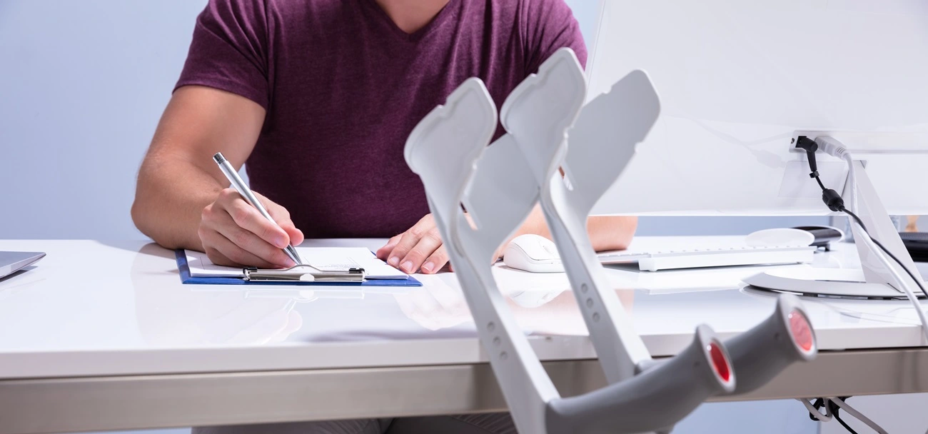Person sitting at desk writing on clipboard near a pair of crutches leaning against the desk.