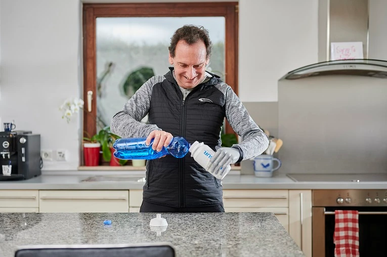 Man in a kitchen pouring water from a blue bottle into a white sports bottle.