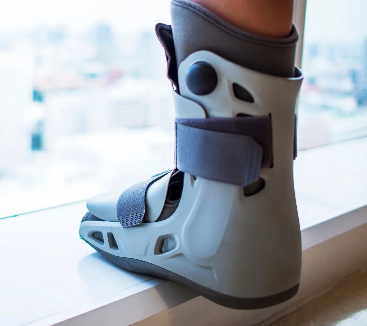 Close-up of a medical walking boot with straps worn on a person's foot near a window.